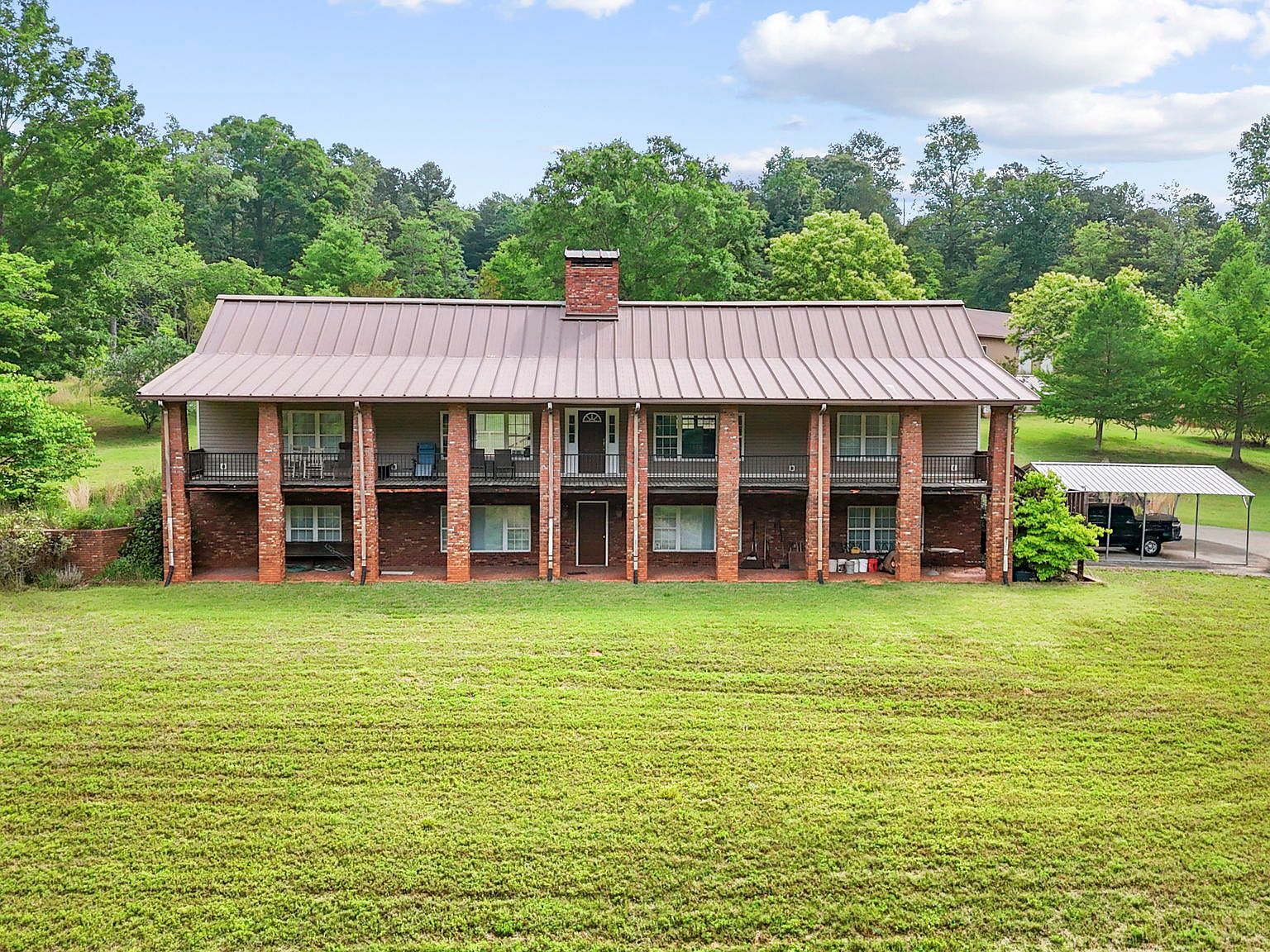Front elevation of the custom-built main residence with red metal roof, brick columns, full-length covered porch, and broad green lawn extending to forest line